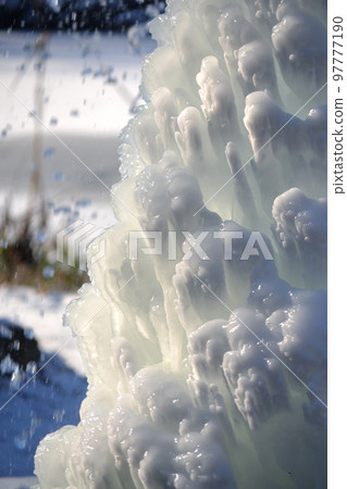 Iced fountain in the park on a winter sunny day. Fountain with frozen water. A jet of water from a fountain that iced over in the cold. Background covered with ice and snow in the park on day 97777190