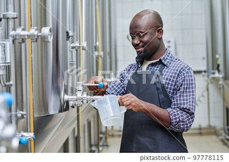 African American worker pouring fresh beer to taste it after brewing while standing in workshop African American worker pouring fresh beer to taste it after brewing while standing in workshop 97778115