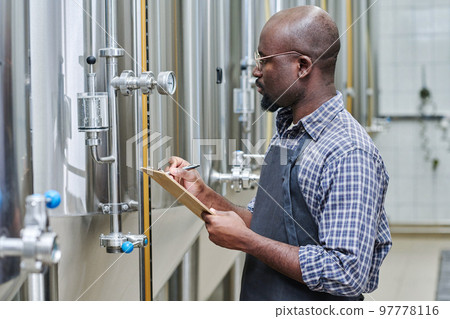 African American worker writing indicators of gauge in document while controlling the work at factory African American worker writing indicators of gauge in document while controlling the work at factory 97778116