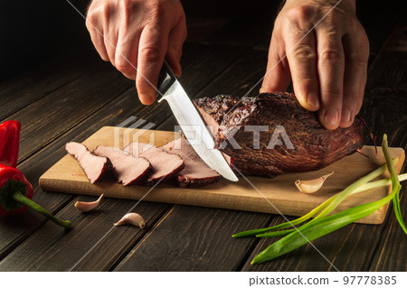 Slicing cooked meat on a kitchen board for dinner. Close-up of a chef hands with a knife. Peasant foods 97778385