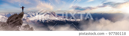 Adventurous Woman Hiker standing on top of icy peak with rocky mountains in background. 97780118