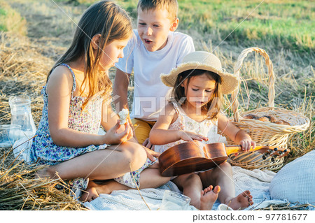 Portrait of glad three kids sitting on blanket in field, playing ukulele, having picnic with milk and tasty bread 97781677