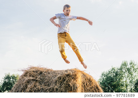 Portrait of barefoot boy in white short and yellow trousers jumping on haystack in field. Outdoor activity. Side view 97781689