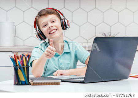 Portrait of young boy wearing black red headphones, sitting at desk in front of laptop, showing thumb up sign gesturing. Portrait of young boy wearing black red headphones, sitting at desk in front of laptop, showing thumb up sign gesturing. 97781767