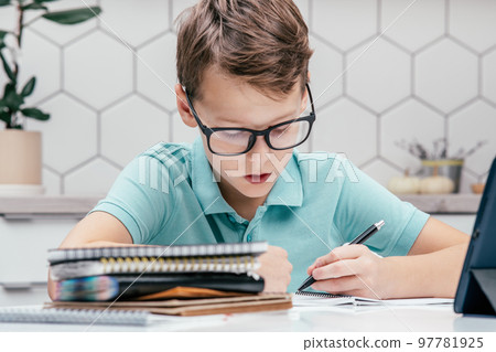 Portrait of preteen focused boy sitting at desk near notebooks, tablet, writing on notebook, holding pen with left hand. 97781925