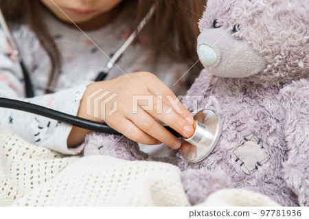 Unrecognizable small girl listen to bear toy patient heartbeat with stethoscope, wrapped in blanket. Little doctor help 97781936
