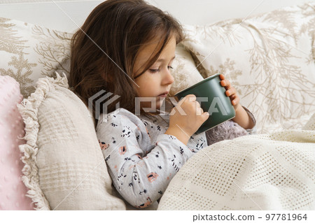 Sweet child, little dark haired girl drink hot unpalatable unpalatable medicine from green mug in bed. Sore throat, pain Sweet child, little dark haired girl drink hot unpalatable unpalatable medicine from green mug in bed. Sore throat, pain 97781964