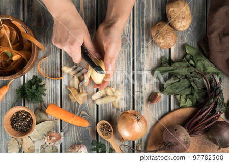 Unrecognizable woman peeling potato with black knife near wooden bowl with carrot peelings little bowl with peppercorns. Unrecognizable woman peeling potato with black knife near wooden bowl with carrot peelings little bowl with peppercorns. 97782500