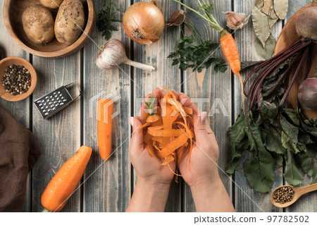 Unrecognizable woman holding carrot peelings in hands near wooden bowl with potatoes, little bowl with peppercorns. Unrecognizable woman holding carrot peelings in hands near wooden bowl with potatoes, little bowl with peppercorns. 97782502