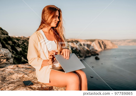 Digital nomad, Business woman working on laptop by the sea. Pretty lady typing on computer by the sea at sunset, makes a business transaction online from a distance. Freelance, remote work on vacation 97789865