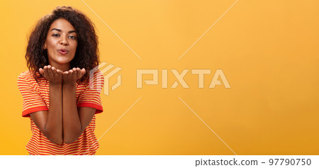 Sending passionate kiss to most loving person. Romantic attractive and stylish young african american girlfriend with curly hairstyle bending towards camera with slight smile, folded lips blowing mwah 97790750