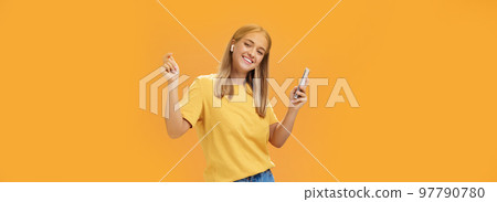 Joyful carefree european woman wearing wireless earbuds holding smartphone dancing with raised hands and pleased happy smile leading vanlife having time to relax and enjoy life over orange wall 97790780