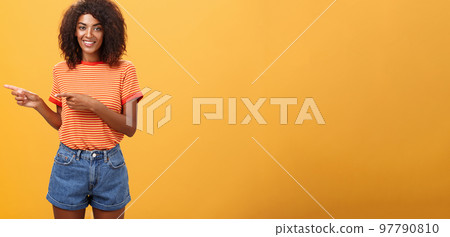 Portrait of charming friendly-looking african-american female coworker in casual outfit pointing left and smiling broadly showing perfect copy space for customers against orange background Portrait of charming friendly-looking african-american female coworker in casual outfit pointing left and smiling broadly showing perfect copy space for customers against orange background 97790810