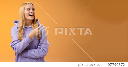 Amused carefree joyful blond european woman in hoodie pointing looking right pleased smiling happily enjoying interesting fascinating peromance making choice in store shopping, orange background 97790872