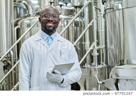 Portrait of successful engineer with digital tablet smiling at camera standing in workshop with big barrels in background Portrait of successful engineer with digital tablet smiling at camera standing in workshop with big barrels in background 97791287