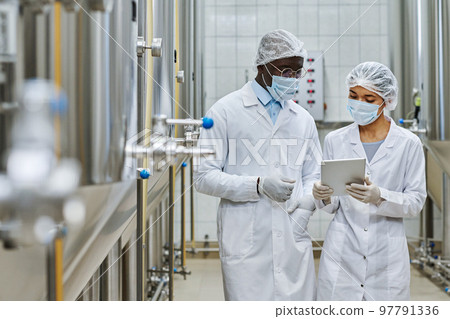 Two professional engineers in white coats and masks discussing something on digital tablet in workshop of brewery Two professional engineers in white coats and masks discussing something on digital tablet in workshop of brewery 97791336