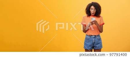 Indoor shot of shocked stunned african american young woman with afro hairstyle staring surprised and excited at smartphone screen holding cellphone reading amazing message over orange wall Indoor shot of shocked stunned african american young woman with afro hairstyle staring surprised and excited at smartphone screen holding cellphone reading amazing message over orange wall 97791400