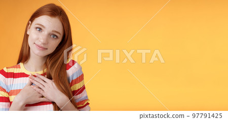 Lovely silly cute redhead young european girl tilting head look gently smiling tender camera press palms heart thankful receive nice gift gazing gratitude happiness, standing orange background 97791425