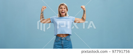 Strong women stand up and fight for rights. Portrait of happy entertained and cool young female athletic blonde with tattoos winking and smiling showing muscles and biceps over blue wall Strong women stand up and fight for rights. Portrait of happy entertained and cool young female athletic blonde with tattoos winking and smiling showing muscles and biceps over blue wall 97791756