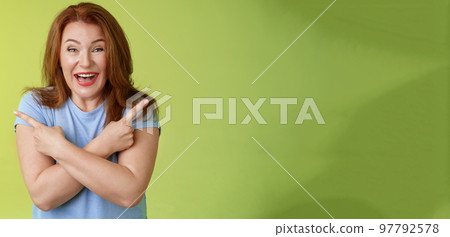 Happy carefree entushiastic redhead funny mature female having fun positive attitude cross arms body pointing sideways show left right products laughing happily like both choices green background 97792578