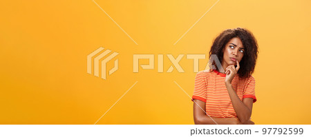 Woman considering to take part in event. Smart focused and creative good-looking african girl with curly hair holding finger on lip gazing concerned left, thinking making decision over orange wall Woman considering to take part in event. Smart focused and creative good-looking african girl with curly hair holding finger on lip gazing concerned left, thinking making decision over orange wall 97792599