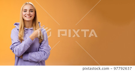Charismatic modern millennial cute blond woman in hoodie smiling happily discussing cool new promo pointing right recommend check out visit interesting store near corner, orange background 97792637