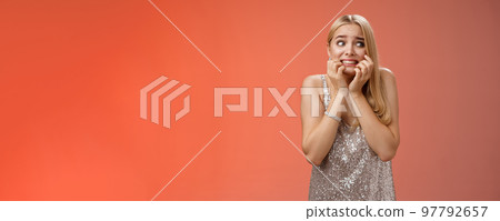 Frightened insecure timid young blond woman afraid feel scared trembling fear stooping biting nails pop eyes right standing horrified red background wear silver evening dress terrified 97792657