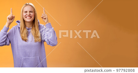 Hopeful charming blond female student praying win first prize achieve dream lifetime hope fortune her side close eyes smiling thrilled pointing up anticipating good news standing orange background 97792808