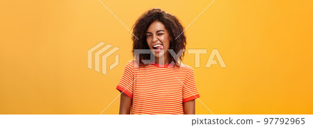 Portrait of daring and emotive confident flirty woman with afro hairstyle winking joyfully showing tongue posing carefree and enthusiastic against orange background flirting with hot guy 97792965