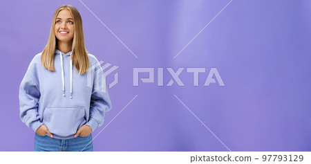 Winter comming. Dreamy kind and cute young girl with straight fair hair and tan wearing hoodie looking at upper left corner with nice white smile and admiring expression holding hand in pockets 97793129