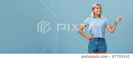 Bossy confident female manager with tattoo on arm holding hand on waist pointing and gazing right with pleased relaxed look giving directions to employees posing over blue background 97793241