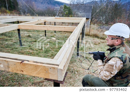 Man building wooden frame house on pile foundation. Worker drilling hole by electric drill in timber framing of future house on construction site. Builder wearing white helmet and protective goggles. 97793402