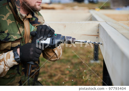Man building wooden frame house on pile foundation. Close up of male worker drilling hole by electric drill in timber framing of future house. Carpentry concept. Man building wooden frame house on pile foundation. Close up of male worker drilling hole by electric drill in timber framing of future house. Carpentry concept. 97793404