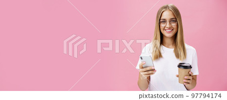 Pleasant friendly-looking girl smiling at camera holding paper cup of coffee and smartphone. Portrait of joyful nice woman drinking morning drink, posing opinion about cafe in internet over pink wall 97794174