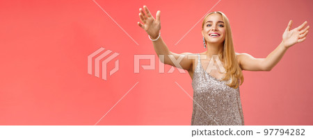 Friendly charming tender hospitable attractive birthday girl in luxury silver dress welcoming guests throw party extend arms cuddle greeting people hug give cuddle smiling broadly, red background 97794282