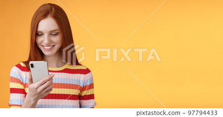 Charming modern redhead girl college student checking message box holding smartphone look happy smiling delighted cellphone display receive hundred likes photo post online, orange background 97794433