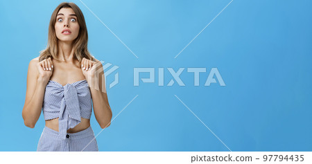 Portrait of stunned speechless and concerned young glamorous female pressing hands to chest popping eyes at camera standing in intense awkward and frightened pose being in stupor against blue wall Portrait of stunned speechless and concerned young glamorous female pressing hands to chest popping eyes at camera standing in intense awkward and frightened pose being in stupor against blue wall 97794435