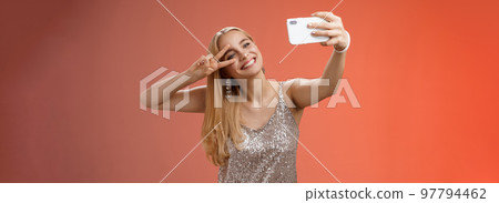 Stylish fabulous glamour young blond woman in glittering silver dress tilting head carefree show peace gesture victory sign extend arm holding smartphone taking selfie recording video post online 97794462