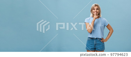 Studio shot of dreamy optimistic good-looking fair-haired female in denim shorts saying shh showing shush gesture with index finger over mouth smiling hiding secret over blue background Studio shot of dreamy optimistic good-looking fair-haired female in denim shorts saying shh showing shush gesture with index finger over mouth smiling hiding secret over blue background 97794623