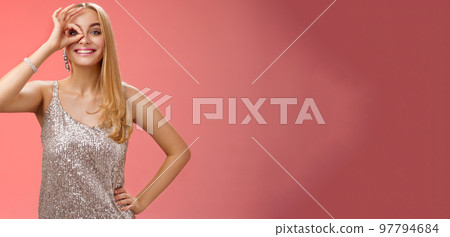 Charming elegant blond european carefree woman in silver glittering evening dress show no problem okay sign look through eye smiling delighted check out awesome promo intrigued, red background 97794684