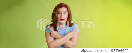 Help me choose. Worried unsure doubtful cute redhead middle-aged silly woman asking advice cross arms body pointing sideways left right look concerned camera make difficult decision green background Help me choose. Worried unsure doubtful cute redhead middle-aged silly woman asking advice cross arms body pointing sideways left right look concerned camera make difficult decision green background 97794695
