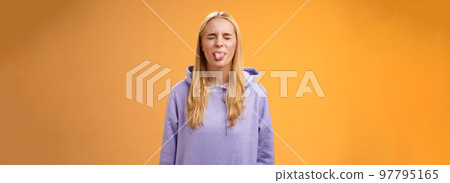 Playful amused charismatic young funny blond girl having fun close eyes showing tongue rebellious fool around unbothered enjoying carefree sunny spring days standing orange background Playful amused charismatic young funny blond girl having fun close eyes showing tongue rebellious fool around unbothered enjoying carefree sunny spring days standing orange background 97795165