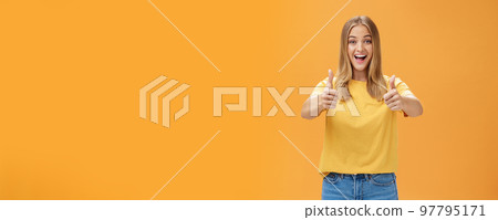 Woman supports with raised thumbs up and amused cheerful smile showing positive attitude expressing like on concept or idea giving approval posing happy and delighted against orange background 97795171