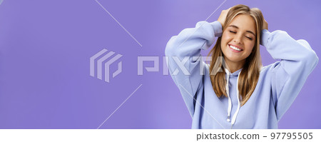 Tender relaxed charming woman with blond hair touching head joyfully smiling broadly with closed eyes daydreaming posing in trendy hoodie against purple wall finally getting rid of acne and pimples 97795505