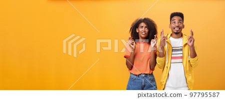 Couple of friends praying and looking up in sky intense crossing fingers for good luck clenching teeth faithfully hoping parents not saw them smoking standing together over orange background Couple of friends praying and looking up in sky intense crossing fingers for good luck clenching teeth faithfully hoping parents not saw them smoking standing together over orange background 97795587