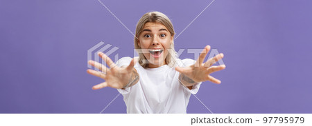 Waist-up shot of amused and excited attractive stylish young woman in white t-shirt pulling hands at camera with desire smiling thrilled and happy wanting hug or take something over purple background 97795979