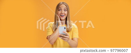 Portrait of surprised and impressed woman reacting to awesome app in smartphone touching cheek from amazement and joy smiling broadly at camera holding cellphone in hand over orange wall 97796007
