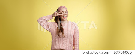 Cheerful happy glamour young european woman in pink knitted sweater, wearing headband, winking flirty and cute, showing victory or pease sign over eye, feeling excited and joyful over yellow wall 97796060