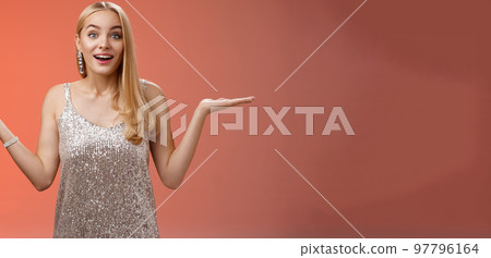 Carefree fascinated young blond girl 20s in silver glittering evening party dress raising hands sideways weighing shrugging smiling amused search right choice, deciding what do red background 97796164