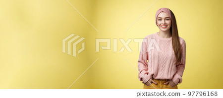 Charming friendly young girl in knitted sweater and headband holding hands in pockets, smiling, looking positive motivated achieving goals having great day, posing positive against yellow background 97796168